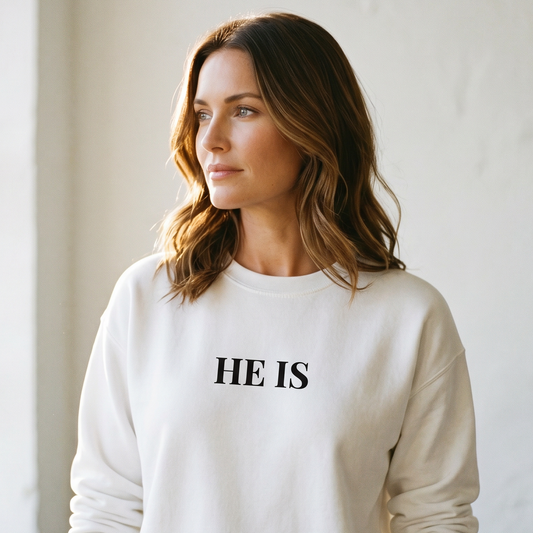 Woman wearing a white sweatshirt with 'HE IS' text in a minimalistic setting