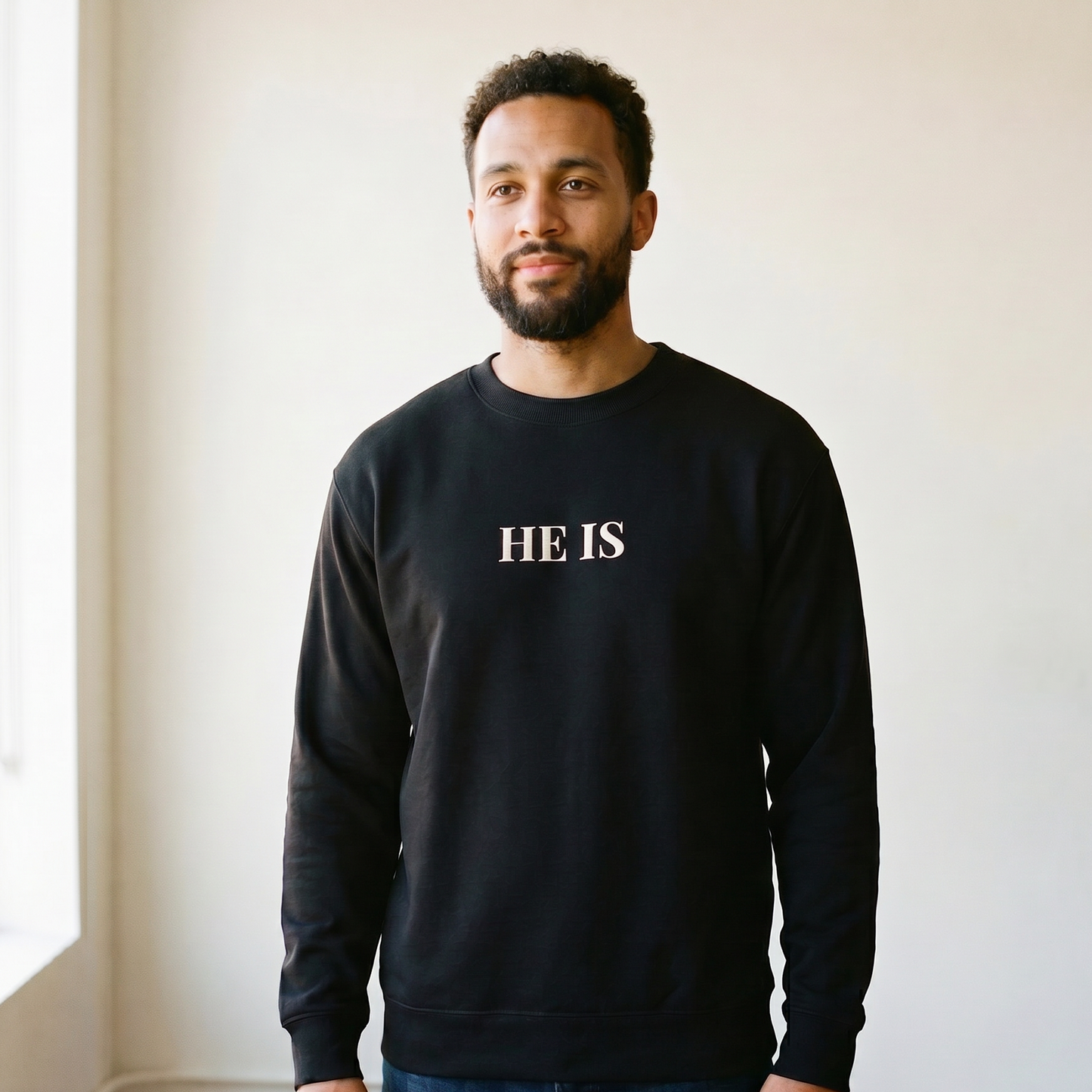 Man wearing a black sweatshirt with 'HE IS' text standing in a room with a window.