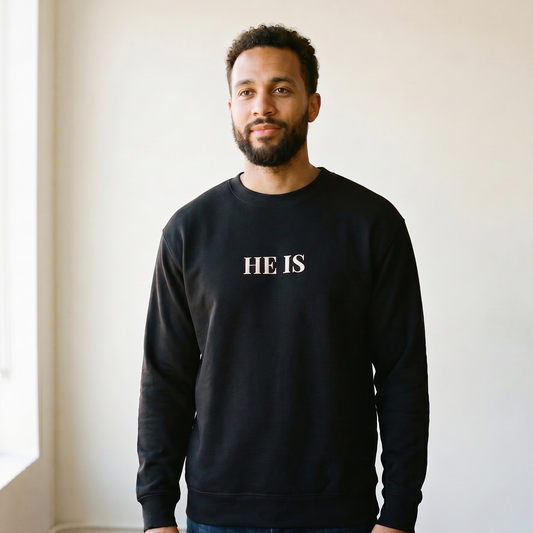 Man wearing a black sweatshirt with 'HE IS' text standing in a room with a window.