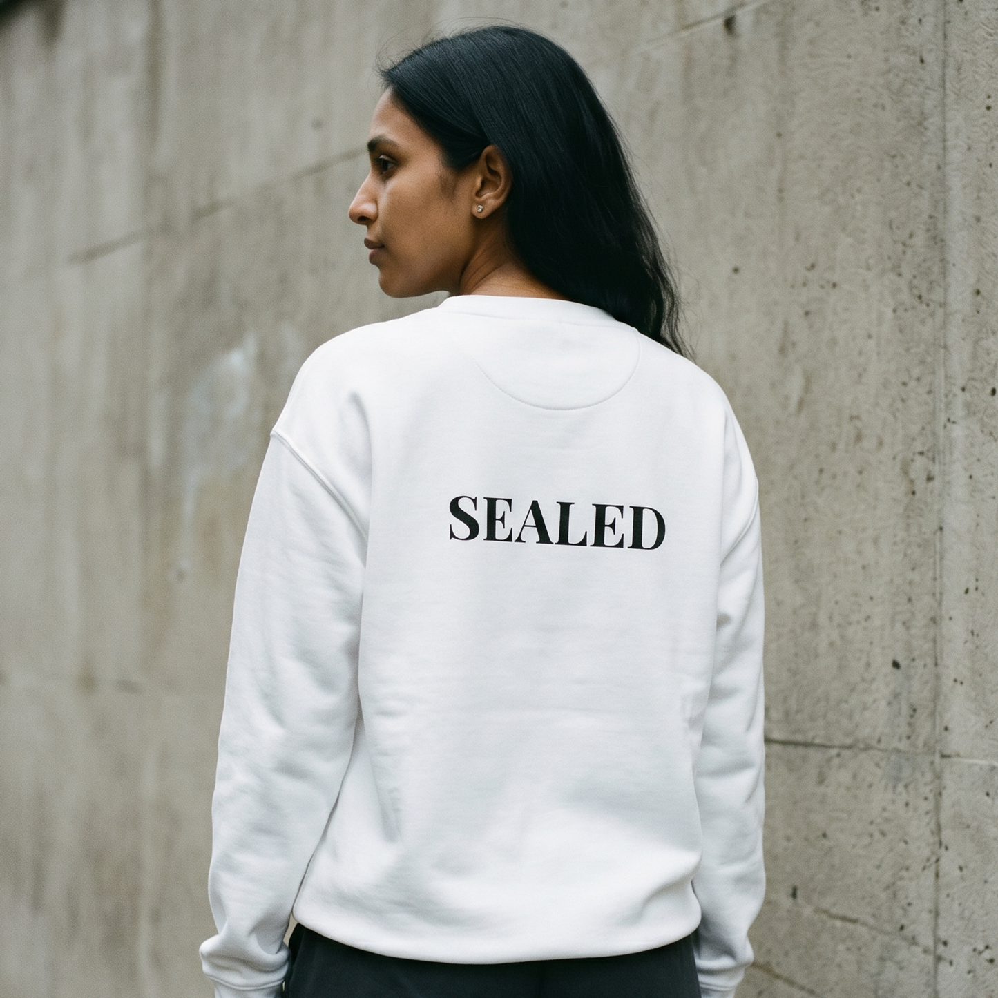 Person wearing a white sweatshirt with 'SEALED' text on the back, standing against a concrete wall.