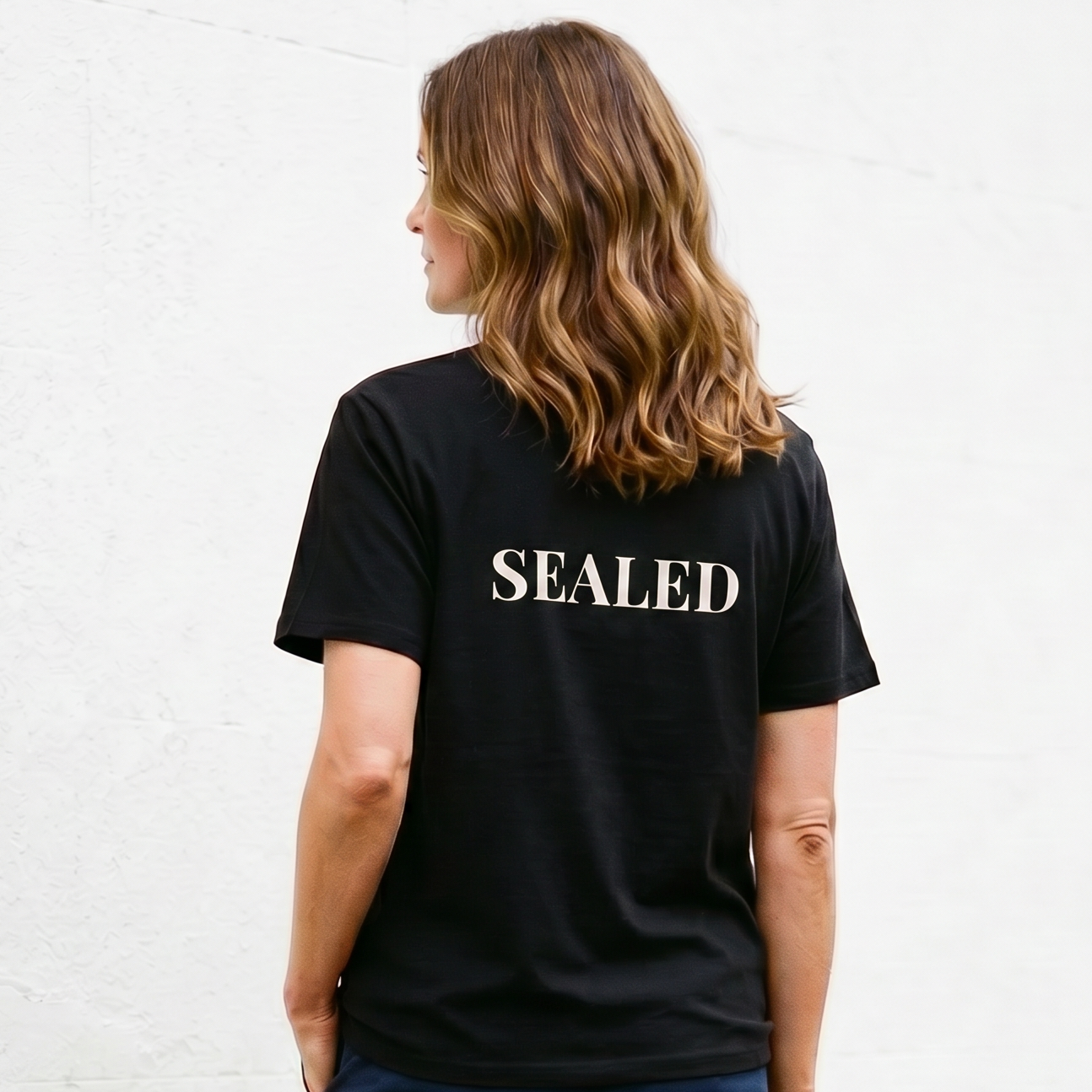 Person wearing a black t-shirt with 'SEALED' on the back, standing against a white wall.