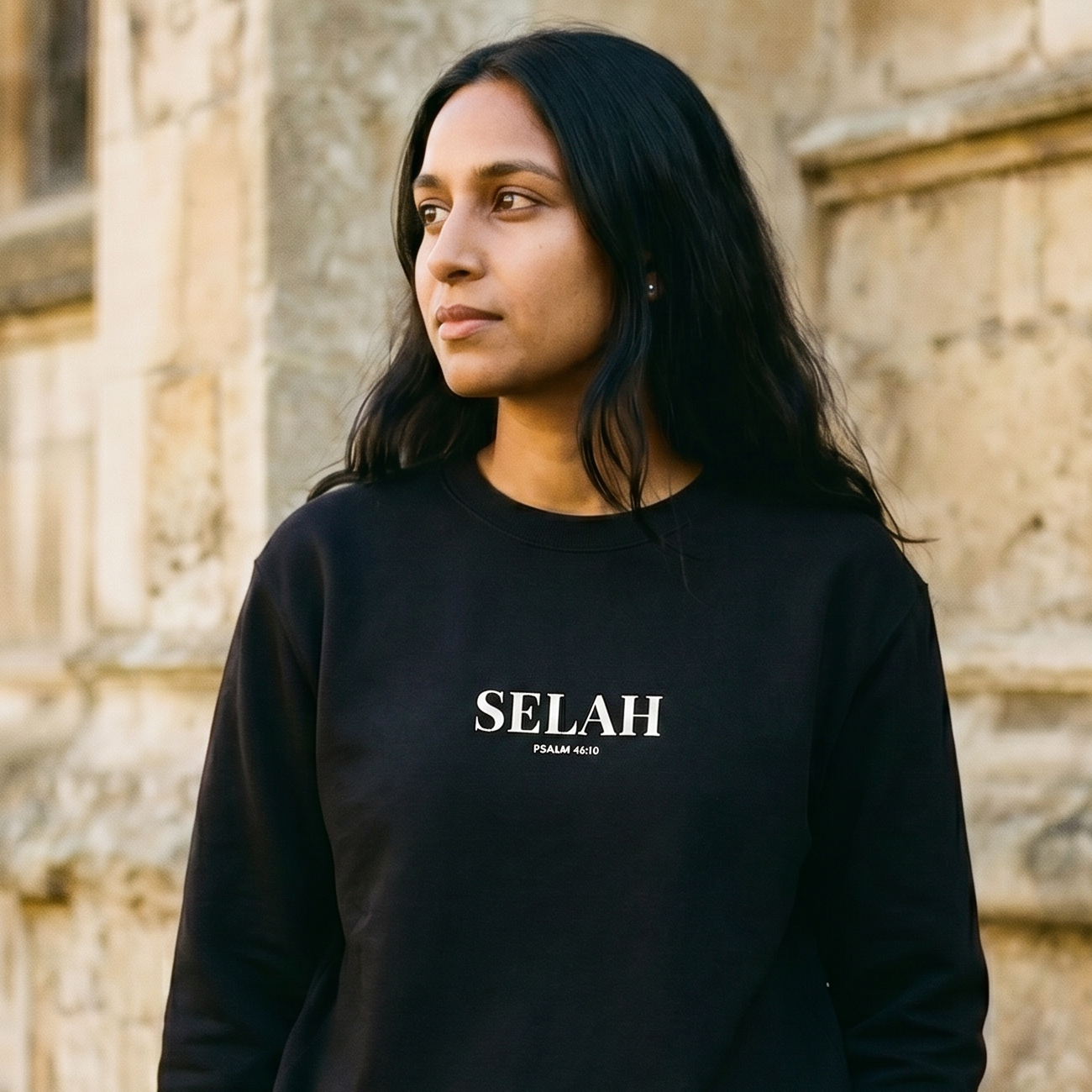 Person wearing a black 'SELAH' sweatshirt standing in front of a stone building.