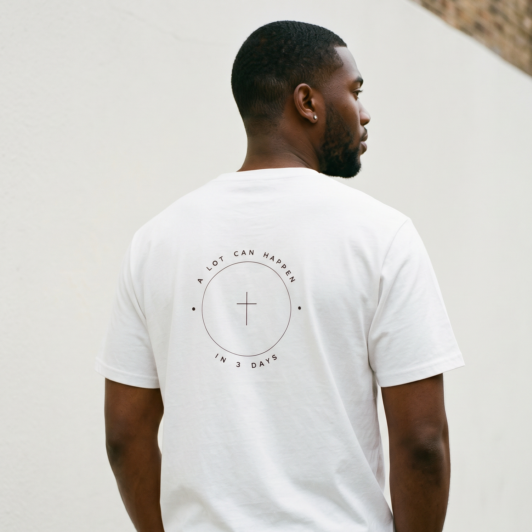 Man wearing a white t-shirt with a circular design and text on the back, standing against a light-colored wall.