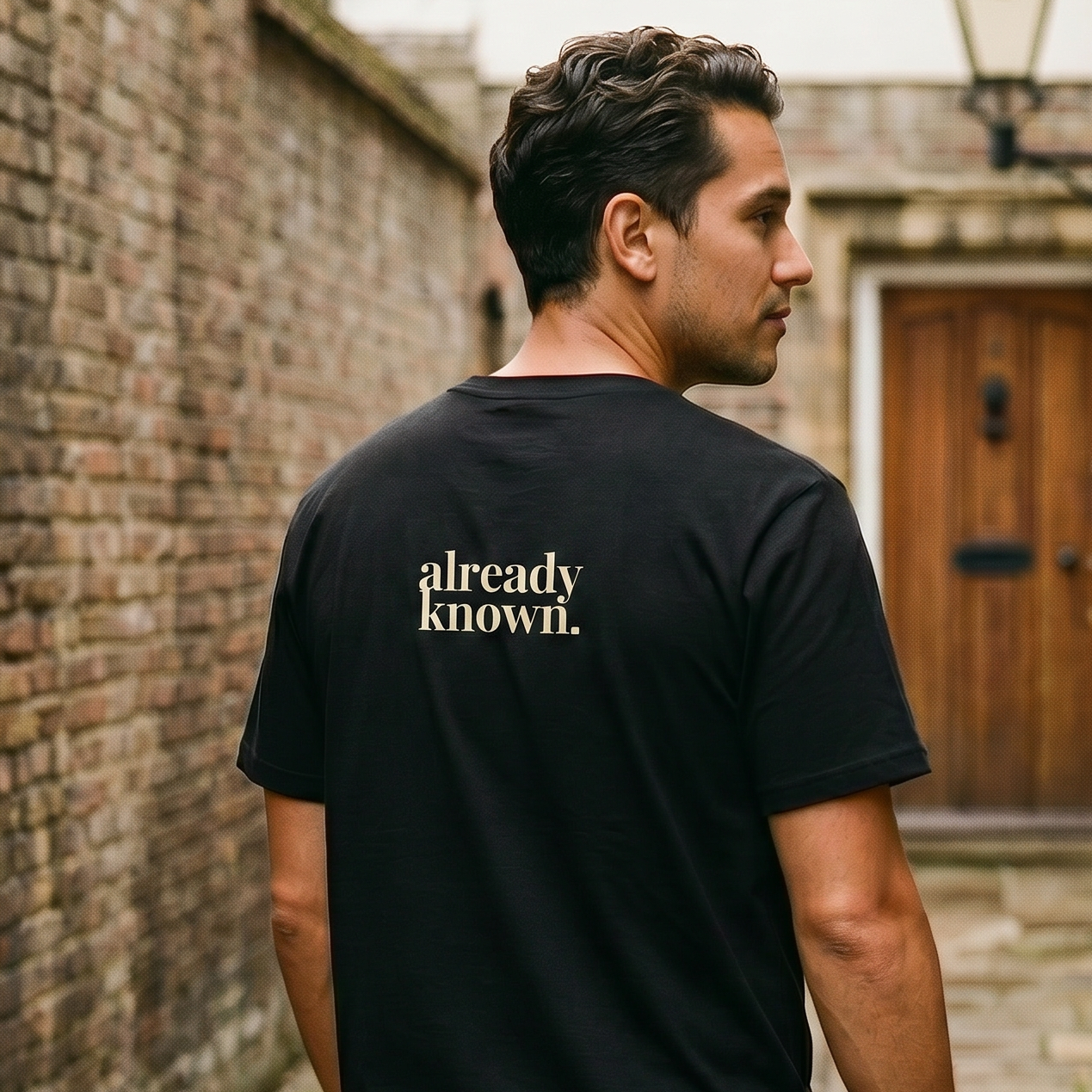 Man wearing a black t-shirt with 'already known' text walking down a narrow alley.