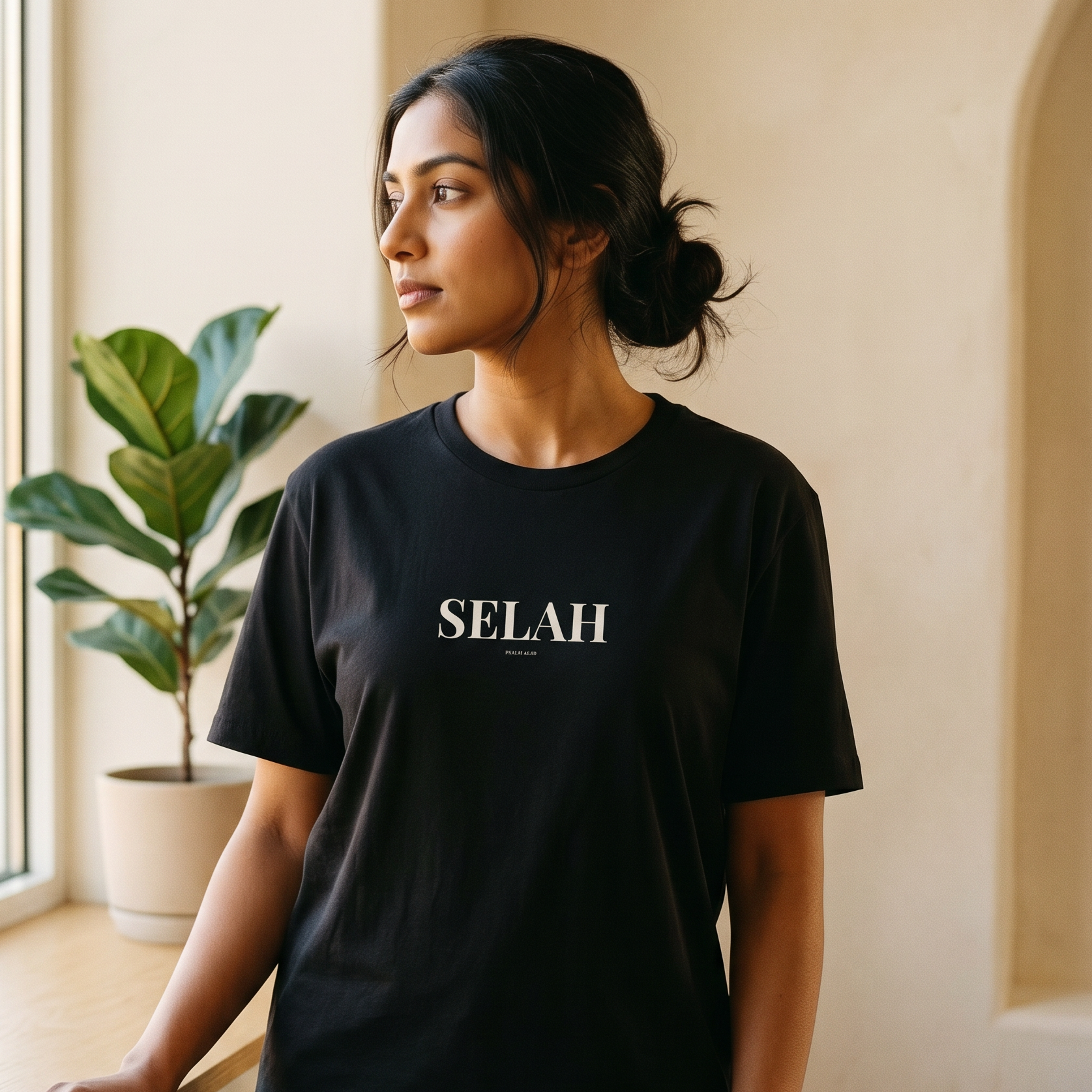 Woman wearing a black t-shirt with 'SELAH' text, standing indoors near a window with a plant.