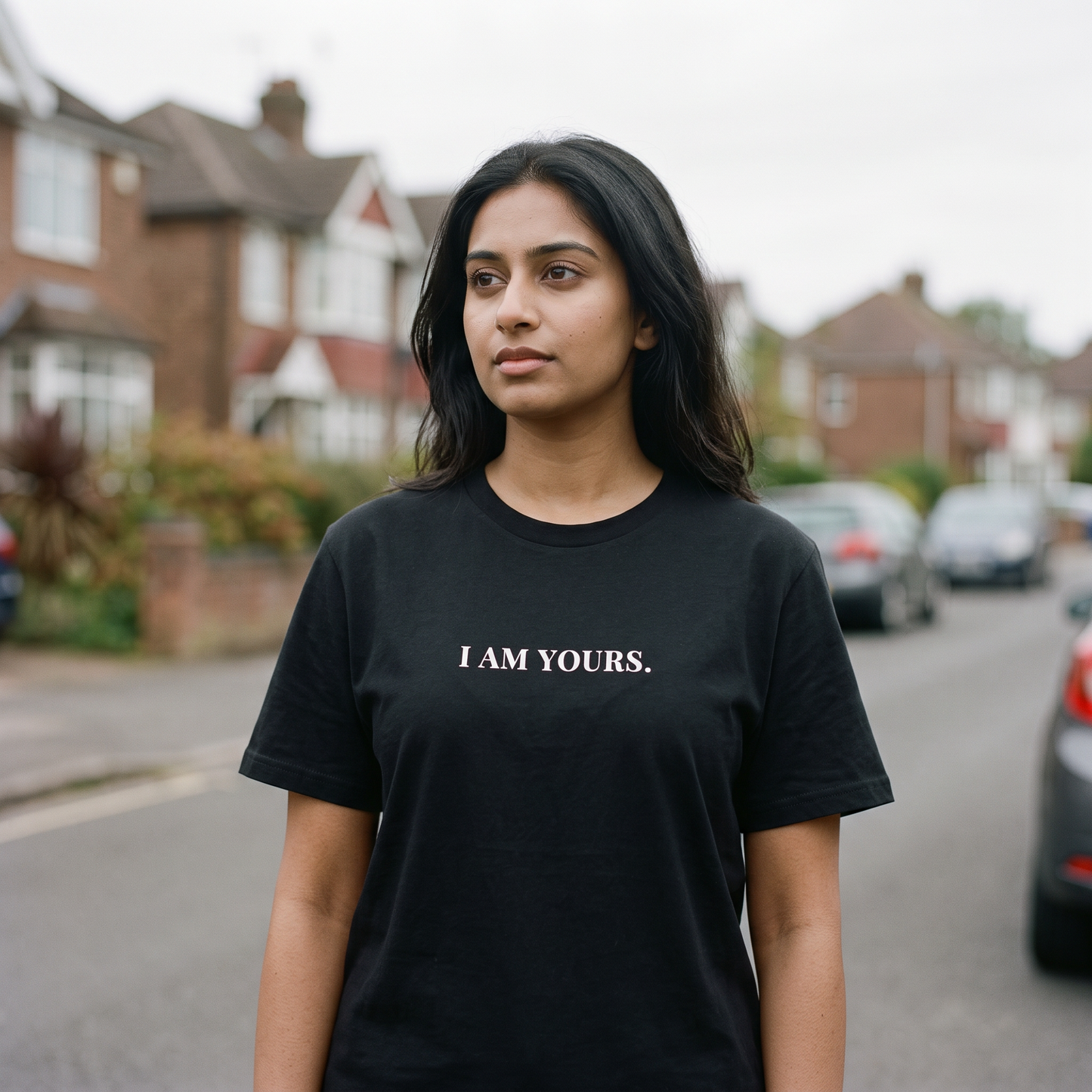 A woman stands on a blurred residential street wearing a black short-sleeve T-shirt with the white printed text 'I AM YOURS.' across the chest.