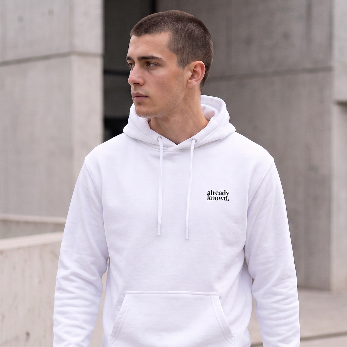 A man wearing a white pullover hoodie with a small black 'already KNOWN' logo on the left chest, standing outdoors in front of a modern concrete building.