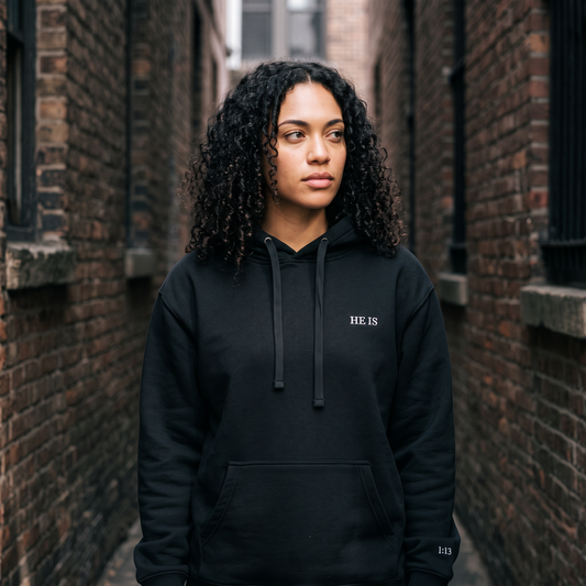 A woman with curly black hair stands in a narrow brick alley wearing a black pullover hoodie with 'HE IS' printed in white on the left chest.