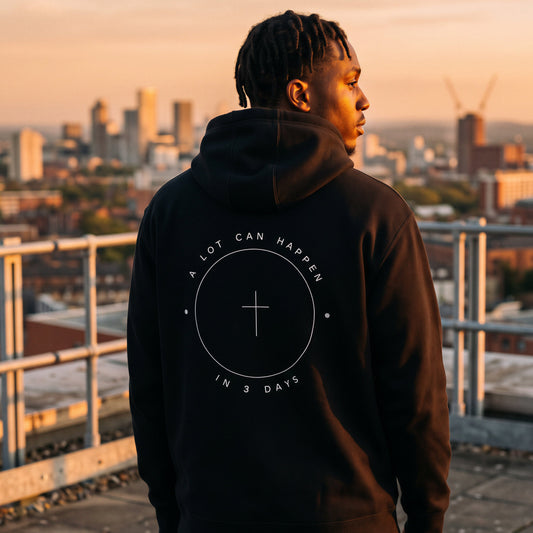 A man stands on a rooftop at sunset wearing a black hoodie with a white circular graphic on the back that reads 'A LOT CAN HAPPEN IN 3 DAYS' around a cross symbol. The city skyline is visible in the background.