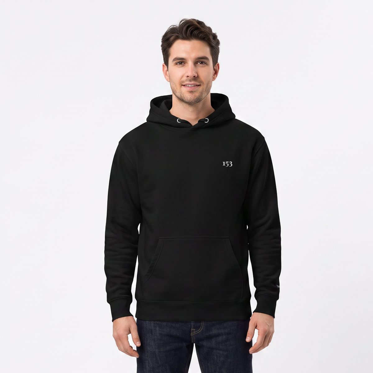 A man wearing a black pullover hoodie with a small white embroidered logo on the left chest, paired with dark blue jeans and white sneakers, standing against a plain white studio background.