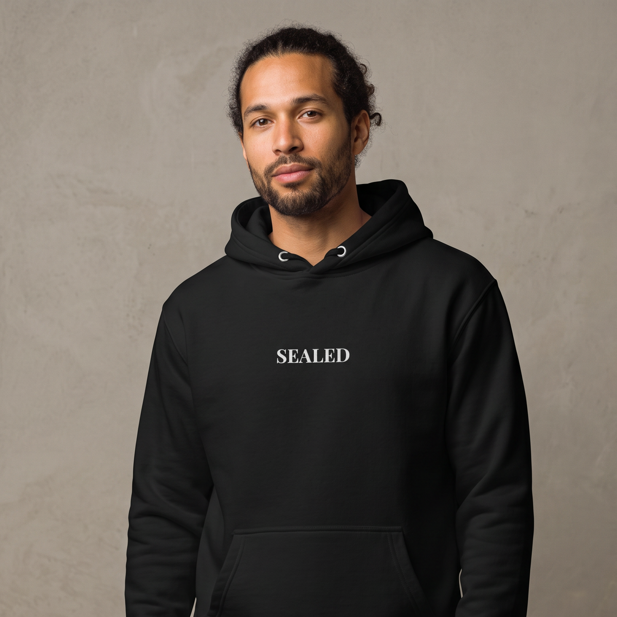 A man wearing a black pullover hoodie with the word 'SEALED' printed in white serif letters across the chest, standing against a textured gray studio background.