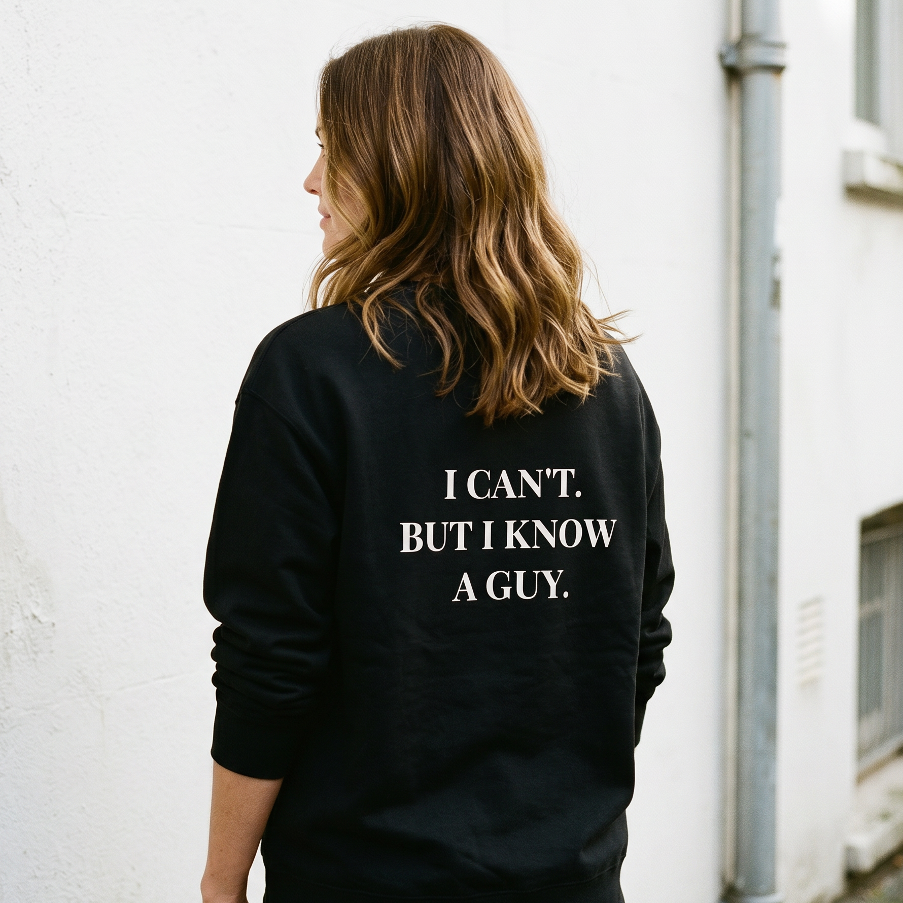 Person wearing a black sweatshirt with text on the back, standing against a white wall.