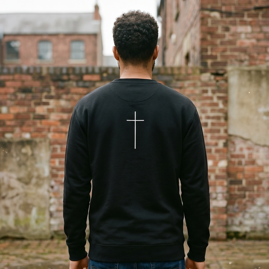 Person wearing a black sweatshirt with a cross on the back, standing in front of a brick wall.
