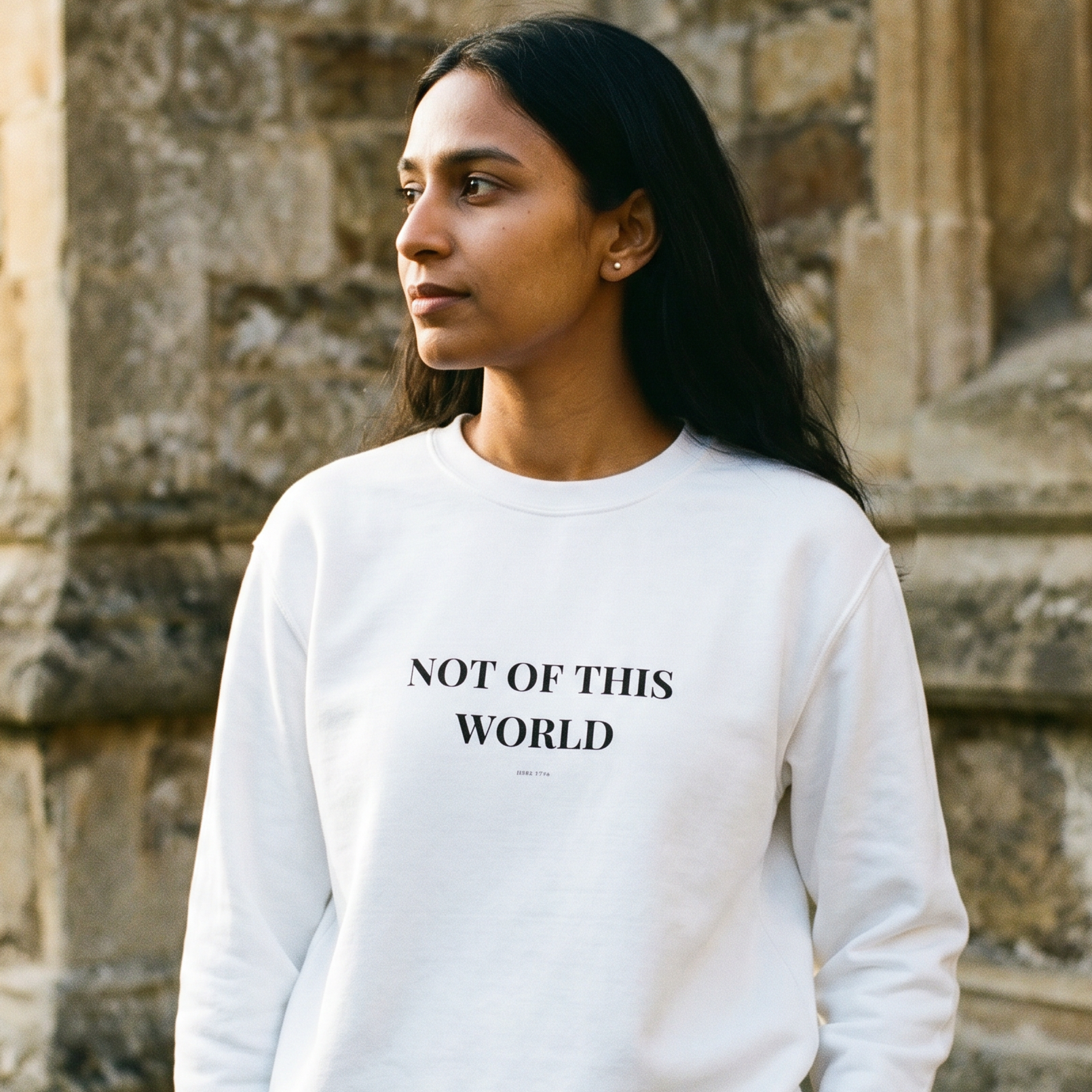 Woman wearing a white sweatshirt with 'NOT OF THIS WORLD' text in front of stone architecture.