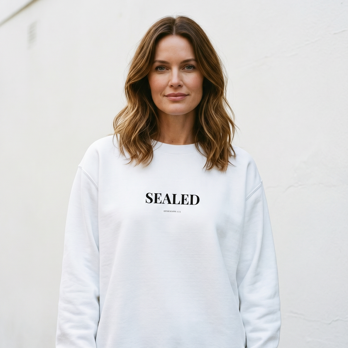 Woman wearing a white sweatshirt with 'SEALED' text against a white wall.