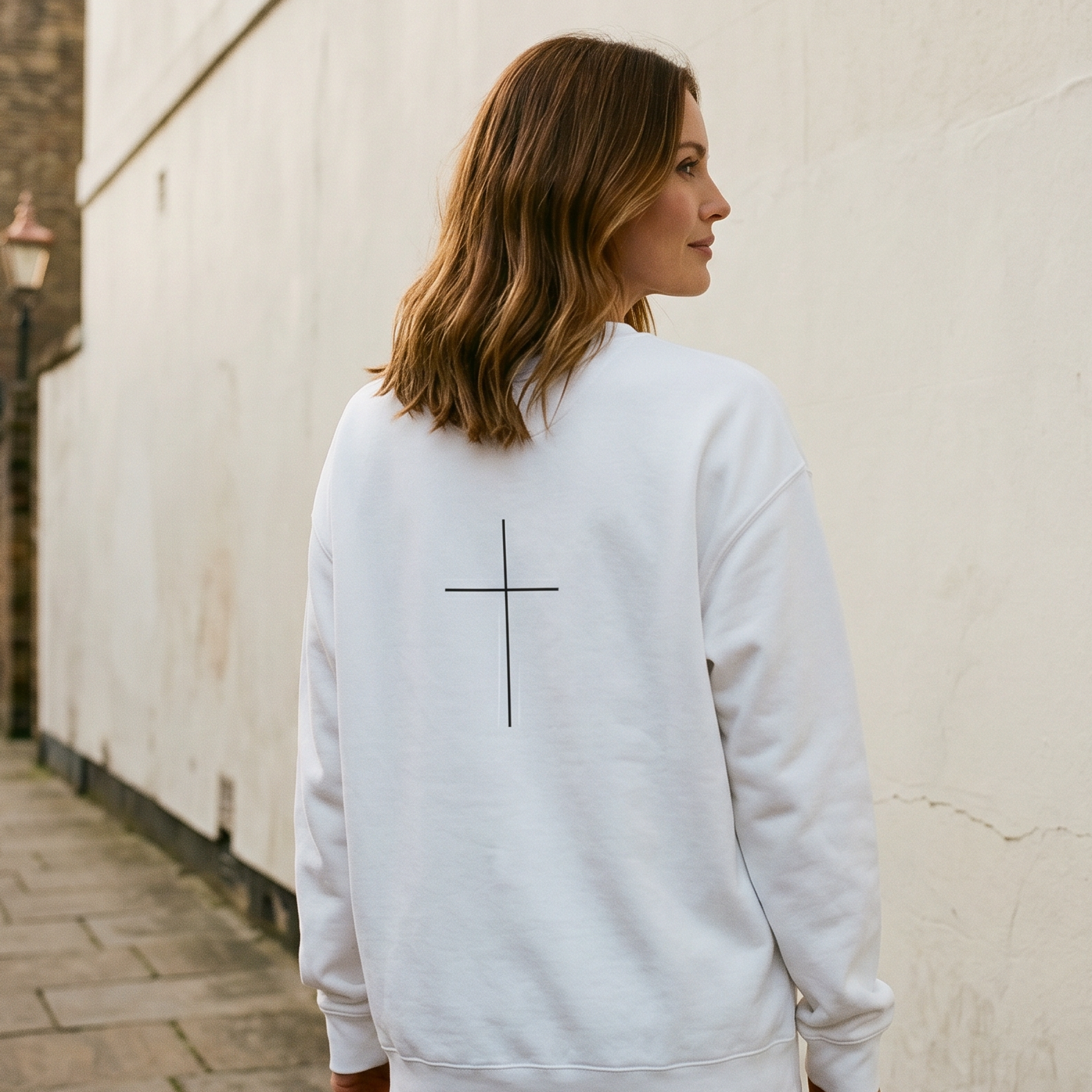 Woman wearing a white sweatshirt with a cross design on the back, standing against a light-colored wall.